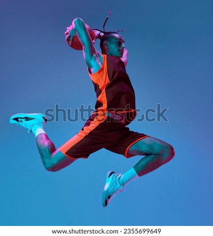 Scoring winning goal. Young man in uniform playing basketball, jumping with ball against blue studio background in neon light. Concept of professional sport, competition, hobby, game, competition