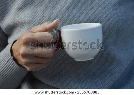 Close-up of female hands holding a white cup. A woman in a gray sweater holds a cup of tea or coffee. Free space for text. Layout.