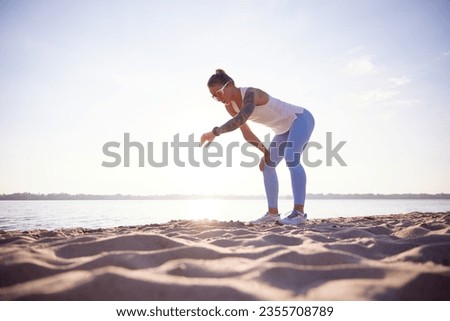 Full-lenght portrait of active woman runner in sport clothes checking athletic tracker on seashore. Woman resting after morning fitness routine. Concept of sport, recreation, healthy lifestyle Royalty-Free Stock Photo #2355708789