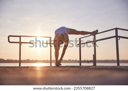 Young woman doing warming-up near seaside. Morning fitness exercise on beach, near the ocean on sunrise. Side legs stretching. Concept of sport, recreation, healthy lifestyle, body care, hobby.