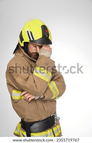 Sad, exhausted firefighter in uniform tiredly rubbing forehead in studio. Side view of fatigued fireman with closed eyes thinking, isolated on gray studio background. Concept of work, emotions.  Royalty-Free Stock Photo #2355719767