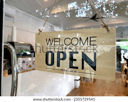 The welcome we are open wooden sign hangs on the glass door at the entrance of the coffee shop.