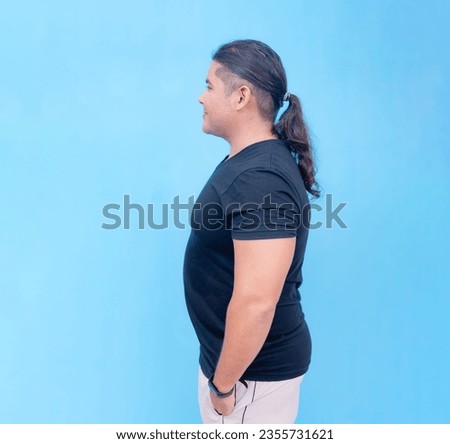 A smiling biracial male standing sideways to the left with his hands inside his pockets. Isolated on a blue background.