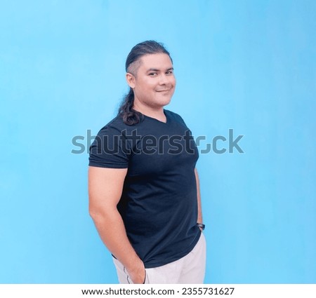 A smiling biracial male standing sideways while looking forwards with his hands inside his pockets. Isolated on a blue background.