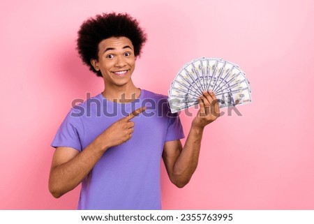 Photo of toothy beaming funny man with afro hairdo dressed blue t-shirt indicating at bunch of dollars isolated on pink color background