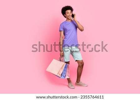 Full size photo of funny positive guy wear stylish t-shirt white pants holding outfit talk on smartphone isolated on pink color background