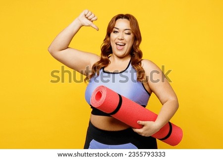 Young chubby overweight plus size big fat fit woman wear blue top warm up training hold yoga mat point thumb finger on herself isolated on plain yellow background studio home gym Workout sport concept