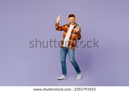 Full body young happy man he wears checkered shirt white t-shirt casual clothes listen to music in headphones use mobile cell phone isolated on plain pastel light purple background. Lifestyle concept