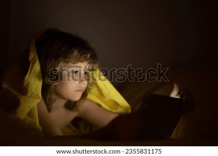 Kid playing on a digital tablet. Kid using tablet for gaming and online learning while lying on the bed, screen light reflex on her face. Child play tablet under blanket at night in a dark room.
