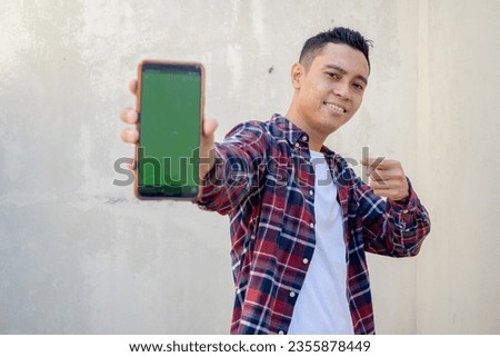 Indonesian man wear casual clothes showing the phone with green screen. The photo is suitable to use for man expression advertising and fashion life style.