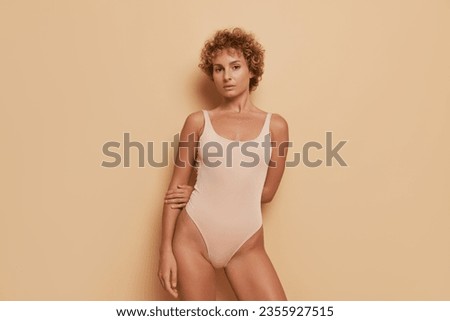 Slim young woman in beige leotard posing on light brown background, keeps hands behind her back, lifestyle concept, copy space