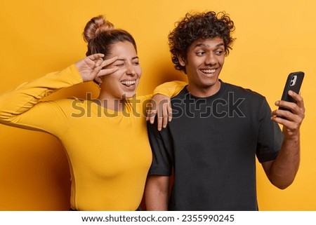 Horizontal shot of positive young woman and man smile gladfully make peace gesture and take selfie concentrated at front camera dressed casually isolated over yellow background make video call