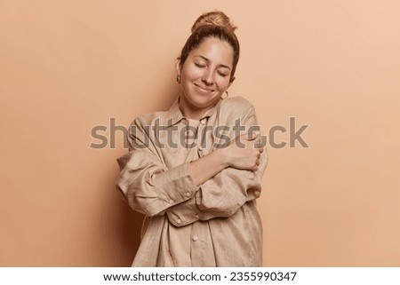 People positive emotions concept. Studio shot of young pretty glad relaxed smiling European lady with closed eyes standing isolated in centre on beige background hugging herself in casual clothes