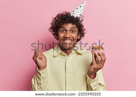 Positive curly haired Hindu man holds small delicious muffin with burning candle clenches fist celebrates birthday in small company of friends wears party hat and yellow shirt isolated over pink wall