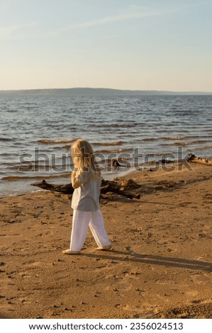 Cute little girl with blonde hair walking barefoot on the beach at sunset on a summer evening. Local travel concept, authentic tourism