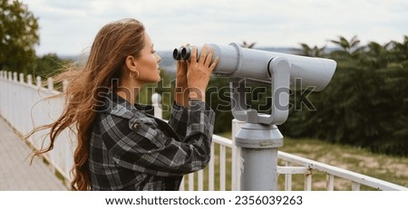 Banner size shot of a young curious woman looking through city binoculars on a windy day. Royalty-Free Stock Photo #2356039263