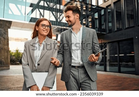Two beautiful successful modern stylish confident smiling adult millennial people walking and talking in business center holding laptop and tablet. Discussing business project strategy at work.