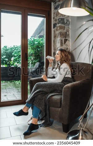 Vertical photo of a young blonde Caucasian woman sending an audio note with the phone in the warmth of her home on the sofa by the window, everyday things at home.