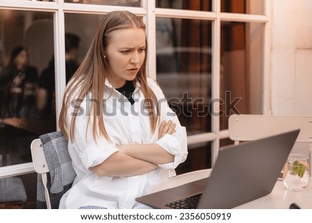 Stressed angry woman talking by video link arguing with colleagues discussing new project sitting at desk in front of laptop computer working from cafe outdoors. Angry woman studying in earphones. Royalty-Free Stock Photo #2356050919