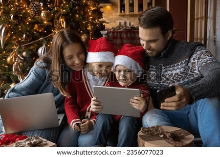 Happy little children siblings watching funny cartoons on digital tablet with affectionate smiling parents, sitting together on floor near decorated Christmas tree, unpacking gifts together at home.