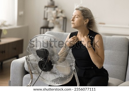 Sick exhausted senior woman with flying blonde hair and closed eyes hair sitting on couch at fresh air blowing from propeller, getting cool, refreshed at fan, feeling overheated, stress relief Royalty-Free Stock Photo #2356077111