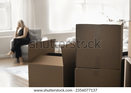 Close up focus on stacked paper boxes for moving, cargo, containers for shipping, transportation. Sad lonely woman in black clothes looking at window in home interior in blurred background