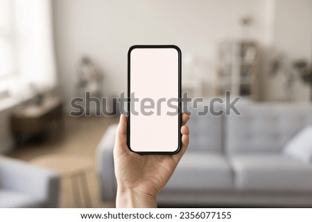 Close up of smartphone in female hand with blank empty screen. Woman holding mobile phone with copy space on display for online smart application interface. Cozy living room interior in background Royalty-Free Stock Photo #2356077155