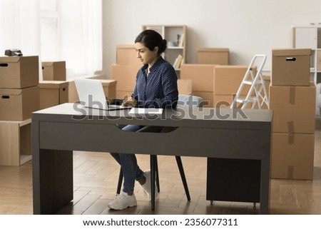 Serious busy young Indian businesswoman working on Internet store project, typing on laptop in storage room among stacked packages, boxes, using e-commerce application for processing order
