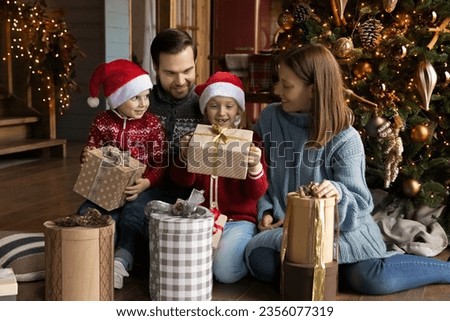 Interested happy little preschool kids boy girl opening wrapped Christmas gifts with affectionate loving couple parents, sitting together on floor near decorated tree, New Year holiday celebration.