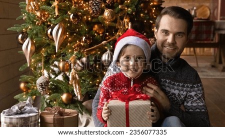 Portrait of smiling bonding young father and little kid son in warm sweater posing near decorated Christmas tree with wrapped gift in hands, feeling excited of New Year holidays celebration at home.