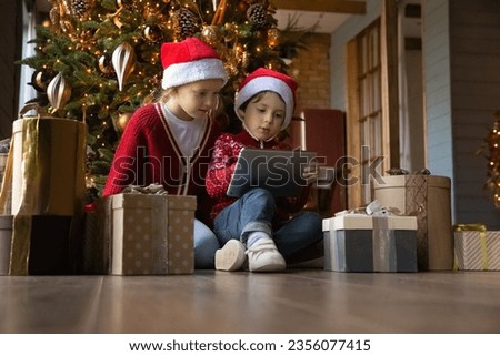 Happy interested little children boy girl in Santa Claus hats sitting on warm heated floor near decorated Christmas trees and gifts, playing online games together on digital tablet, tech addiction.