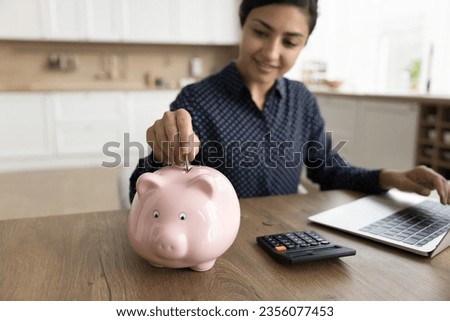 Positive young Indian freelance business woman saving money for future, dropping coin into pink piggy bank on workplace table at home, enjoying financial management, investment