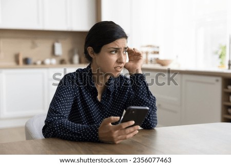 Concerned young Indian woman holding smartphone, looking away in deep thoughts, thinking on problems, feeling doubt, uncertain, worried, anxious, touching head, chatting at home table Royalty-Free Stock Photo #2356077463