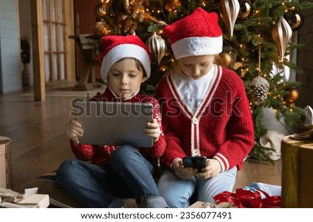 Happy small kids boy girl sitting near decorated Christmas tree enjoying using different electronic gadget, playing games or watching videos in social network on winter holidays, tech addiction.