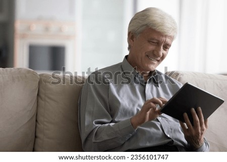 Cheerful retired pensioner man using online app on digital tablet on home sofa, enjoying Internet communication, touching touchscreen, laughing, smiling, relaxing, enjoying leisure