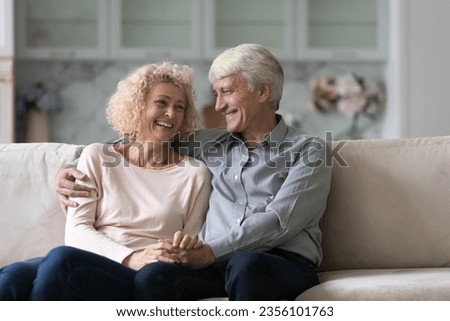 Joyful older retired husband and wife enjoying dating, love relationship, anniversary, leisure together, hugging, holding hands on home couch, talking, smiling, laughing at joke Royalty-Free Stock Photo #2356101763