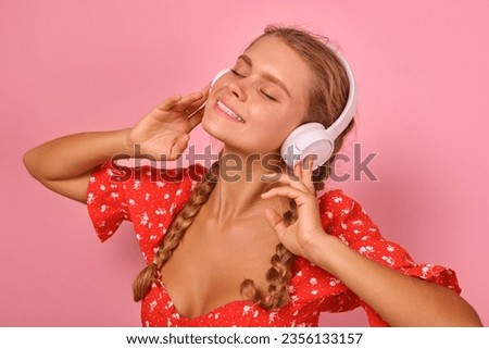 Young positive pretty Caucasian woman presses headphones to ears and smiles enjoying voice of favorite music artist and imagining dance with boyfriend stands posing in pink studio.