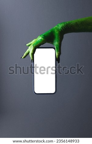 Vertical image of green monster hand holding smartphone with copy space on grey background. Halloween, tradition and celebration concept. Royalty-Free Stock Photo #2356148933