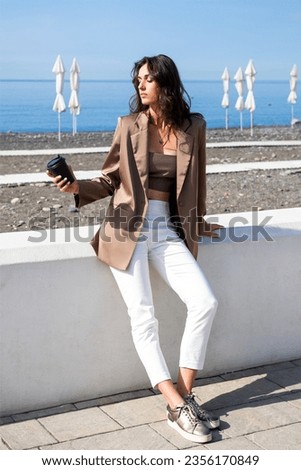 tall, slender brunette model is sitting on the sea embankment. A full-length portrait of beautiful woman in brown jacket, top and white trousers against the background of the sea