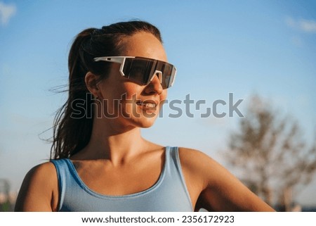 Close up portrait of caucasian young woman in sportswear and protective sunglasses looks away at sunset time relaxing after jogging, cycling. Purposeful sporty girl enjoying fresh air outdoors. Health Royalty-Free Stock Photo #2356172923