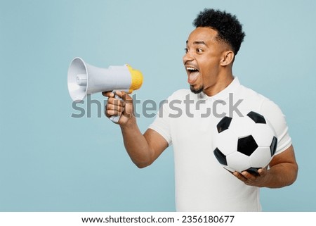 Young man fan wear t-shirt scream in megaphone announces discounts Hurry up cheer up support football sport team hold in hand soccer ball watch tv live stream isolated on plain blue color background