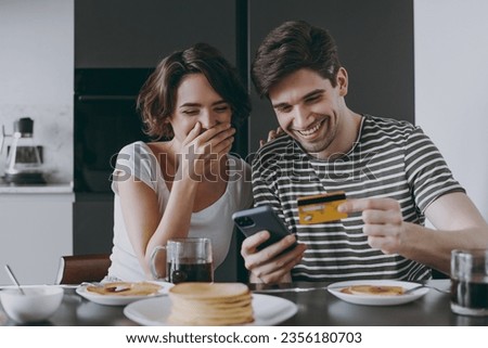 Young happy couple two woman man in casual clothes sit by table eat pancake use mobile cell phone credit bank card shop online order delivery in light kitchen at home together Healthy diet concept. Royalty-Free Stock Photo #2356180703