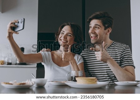 Young happy couple two woman man 20s in casual clothes sit by table eat pancake do selfie by mobile cell phone show thumb up gesture cook food in light kitchen at home together Healthy diet concept