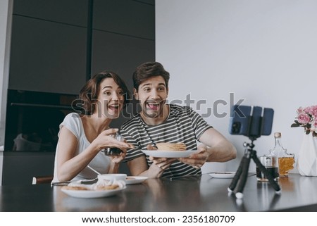 Young fun couple two woman man in casual clothes sit by table eat show pancake talk video call mobile cell phone drink drinking coffee cook food in light kitchen at home together Healthy diet concept