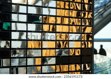 Wall made of polished metal mirrored rectangles imitating brickwork. Interior element and silhouette of people in the background. Photo. Selective focus. Daylight. Close-up