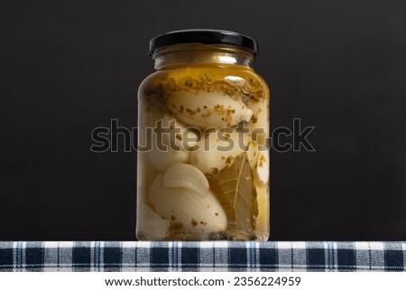 Photograph of sealed jar of pickled onions on a table with a checkered cloth, on dark background.