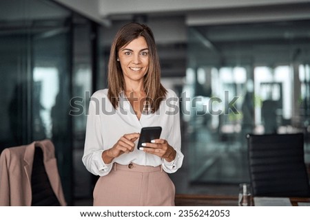Mid aged business woman holding cellphone standing in office, portrait. Mature 40 years old professional businesswoman manager executive using mobile phone working on smartphone at workplace. Royalty-Free Stock Photo #2356242053
