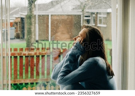 Thoughtful sad woman sitting on a windowsill with a reflection on a window. Mental health and seasonal disorder due to rainy cold weather, end of summer concept Royalty-Free Stock Photo #2356327659