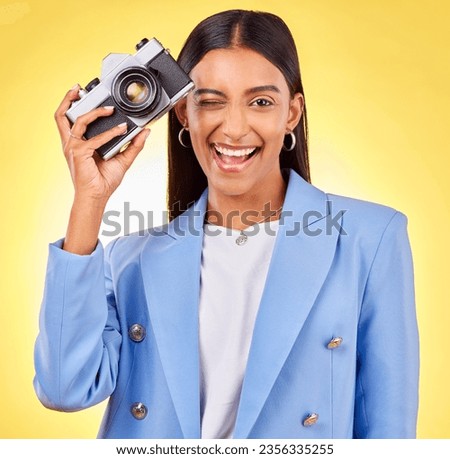 Camera, photography and woman portrait with a smile and wink for picture in studio. Happy, female person and yellow background with creative, memory and funny emoji face with photographer skill