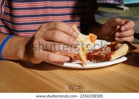 Boy eating pizza with hands in outside restaurant during sunny day. He has red t shirt with straps and blue rubber bracelet on his right hand. Royalty-Free Stock Photo #2356338585